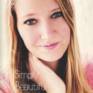 Könnte beinhalten: Ein Porträt einer Person mit langem blonden Haar und blauen Augen, die ein rosafarbenes, strukturiertes Kleidungsstück trägt. Der Text Simply Beautiful ist in Weiß in der unteren linken Ecke des Bildes gedruckt, mit klarer Sicht auf Gesicht und Schultern.
