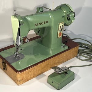 May include: A vintage mint green Singer sewing machine with a chrome needle bar and presser foot, mounted on a rectangular wooden base. The machine includes a matching mint green foot pedal connected by a long electrical cord and plug.