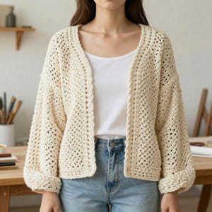 May include: Cream-colored, open-front crochet cardigan with long sleeves. The cardigan has a loose, boxy fit and a textured, open weave design. The sleeves are cuffed at the wrists. The cardigan is worn over a white top and blue jeans.