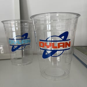 May include: Two clear plastic cups with the name "DYLAN" printed on them. The text is in orange and blue, with a stylized design. The cups are empty and appear to be disposable.