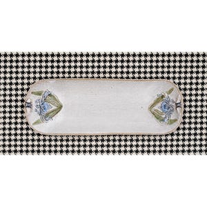 May include: An elongated, oval-shaped ceramic tray with a textured, off-white finish. The tray features symmetrical floral motifs on both ends, displaying blue petals and green leaves, with a slightly raised rim and a smooth, flat central surface.