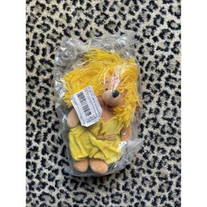 May include: A plush lion toy with yellow yarn hair and a yellow dress, packaged in a clear plastic bag. The lion has a brown nose and is wearing a gold-colored belt. A barcode label is attached to the bag.