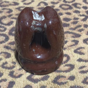 Pode incluir: Um isolador elétrico vintage castanho escuro com acabamento brilhante. A peça de cerâmica tem uma forma única com um orifício central e uma superfície texturizada. Algumas áreas mostram sinais de desgaste.