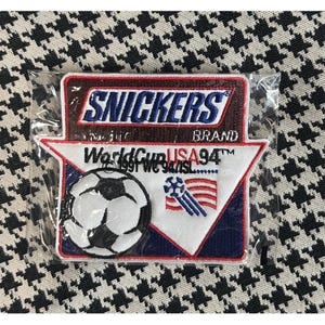 Pode incluir: Um patch da marca Snickers com o texto "World Cup USA 94", uma bola de futebol e a bandeira americana. O patch está em uma embalagem plástica transparente e sobre um fundo com estampa em preto e branco.