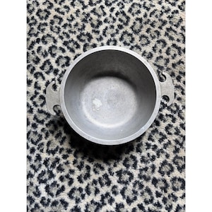 May include: A round, silver-colored cooking pot with two handles. The pot is made of metal and has a simple, utilitarian design. The pot is empty and appears to be clean. The background is a patterned rug.