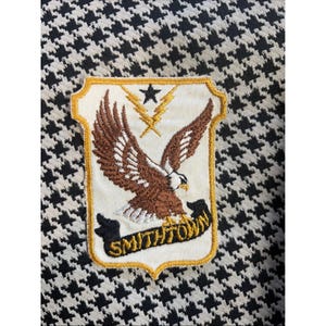 Puede incluir: Parche bordado con un águila marrón y blanca con las alas extendidas, una estrella negra y rayos. El parche está bordeado en dorado y tiene la palabra "Smithtown" en una pancarta negra. El fondo es un estampado de pata de gallo en blanco y negro.