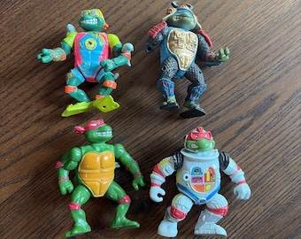 Pode incluir: Quatro figuras de ação das Tartarugas Ninja. As figuras são de várias cores, incluindo verde, vermelho, azul e amarelo. Cada figura tem uma armadura e acessórios únicos, com expressões faciais e poses distintas. As figuras estão sobre uma superfície de madeira castanha.