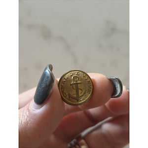 May include: A gold-colored button with an anchor design in the center. The button has a circular shape with a detailed border. The word "CANADA" is visible around the edge of the button. The button is held between two fingers.