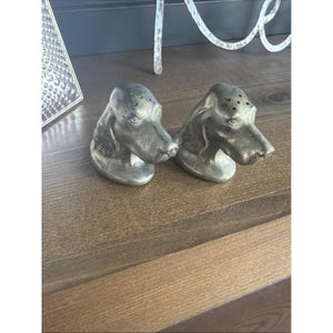 May include: A pair of metallic silver-toned dog head salt and pepper shakers. Each shaker features a sculpted canine head with long ears and a series of small holes on the top of the head for dispensing seasoning. The finish has a subtle sheen.