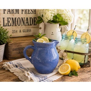 May include: A blue ceramic pitcher filled with lemonade, garnished with lemon slices and mint. Two mason jars of lemonade with lemon slices and striped straws sit in a metal tray. A wooden sign reads "FARM FRESH LEMONADE 25¢ A GLASS."