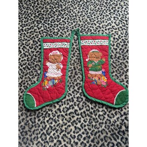 May include: Two quilted red Christmas stockings with green trim and a white cuff featuring a holly pattern. Each stocking displays a brown teddy bear illustration, one wearing a nightgown and the other in pyjamas, surrounded by colourful gift boxes.