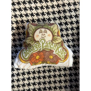 May include: A vintage Cabbage Patch Kids doll with a smiling face, surrounded by green leaves. The doll has the Cabbage Patch Kids logo and floral accents. The doll is on a black and white houndstooth patterned surface.