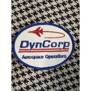 May include: An oval patch with the DynCorp Aerospace Operations logo. The patch features a white background with a blue border. The DynCorp text is in blue, and the Aerospace Operations text is in red. A red airplane graphic is above the DynCorp text.