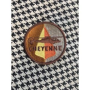 Puede incluir: Un parche redondo con un diseño de helicóptero marrón y la palabra "CHEYENNE" en texto marrón. El parche tiene un borde marrón y un fondo marrón, amarillo y naranja. Se encuentra sobre un patrón de pata de gallo en blanco y negro.