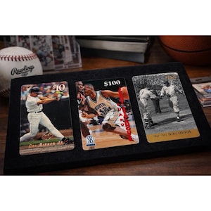 May include: Three sports trading cards displayed on a black felt surface. The cards feature images of baseball and basketball players. One card shows Cal Ripken Jr. and is priced at $10. Another card features a basketball player and is priced at $100.
