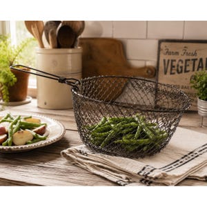 May include: A dark metal wire mesh basket with a long handle, containing a pile of fresh green beans. The basket features a fine, diamond-patterned wire construction and is positioned on a folded, light-colored fabric cloth.