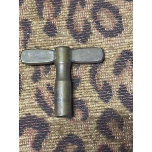 May include: A vintage brass clock key with a T-shaped design. The key has a cylindrical body with two rectangular wings on top. The key is likely used to wind a mechanical clock.