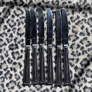 Op de afbeelding: Zes roestvrijstalen dinermessen met een reflecterende afwerking. De messen hebben een eenvoudig, elegant ontwerp met een lang, slank lemmet en een handvat met verticale groeven. De messen zijn gerangschikt op een patroon.