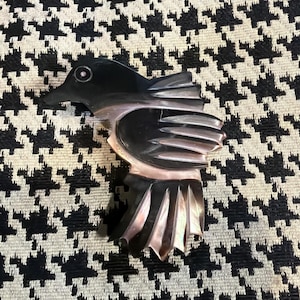 May include: A bird-shaped brooch crafted from polished dark material with iridescent highlights. The piece features a carved head with a circular eye detail, a wing with layered segments, and a fanned tail, showcasing smooth, reflective surfaces.
