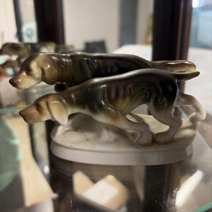 May include: A ceramic figurine featuring two dogs in a hunting pose. The dogs have brown and black markings with tan ears and snouts. The figurine is mounted on a white oval base, showcasing a classic hunting theme.