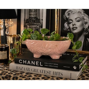Può includere: Una fioriera in ceramica rosa con un motivo a foglie, piena di piante verdi, poggia su due libri. I libri sono intitolati "CHANEL COLLECTIONS AND CREATIONS" e "BAZAAR GREATEST HITS". Sono visibili una lampada nera e un portacandele dorato.