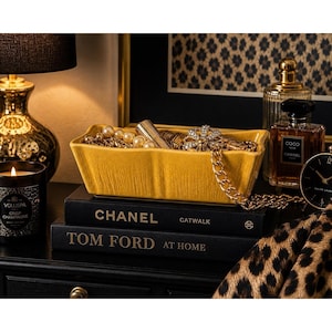 May include: A yellow rectangular jewelry dish filled with gold necklaces, pearls, and a lipstick. The dish sits atop two books, "Chanel Catwalk" and "Tom Ford at Home." A black candle and perfume bottles are also visible.