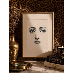 May include: A framed black and white print of a woman's face. The print is in a gold frame and is displayed on a marble surface. A gold vase with dried flowers, a magnifying glass, and books are also on the surface.