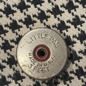 Puede incluir: Un flexómetro metálico vintage con la inscripción "LITTLE PAL", "MADE IN U.S.A." y "3 FEET". El flexómetro tiene un centro rojo y está colocado sobre un estampado de pata de gallo en blanco y negro. 3 pies son aproximadamente 91 cm.