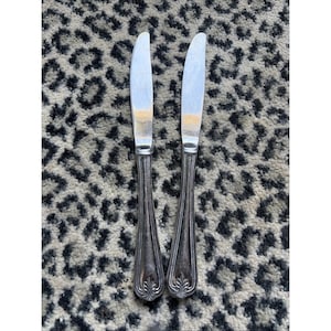 May include: Two silver butter knives with a decorative handle. The blades are shiny and the handles have a detailed, ornate design. The knives are placed on a patterned surface.