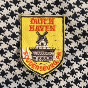 May include: A yellow patch with the words "DUTCH HAVEN" and "SOUDERBURG PA" in orange and white lettering. The patch features a windmill illustration. The background is a black and white houndstooth pattern.