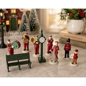 May include: A collection of miniature ceramic figurines featuring a green park bench, a street clock, and several figures including musicians in red uniforms, a person in a red coat, a guard in a black bearskin hat, and a newsboy holding a paper.