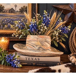 May include: A ceramic hat-shaped planter with a marbled tan, brown, and grey finish, filled with a bouquet of blue lupine flowers, white filler flowers, and stalks of wheat, resting on two stacked books labeled TEXAS and RANCH HOUSE.