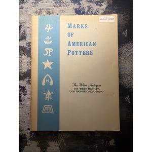May include: A softcover book titled Marks of American Potters with a light blue vertical stripe on the left featuring white symbols including an anchor, star, and fleur-de-lis. The cover includes the text The Weirs Antiques, 101 West Main St.