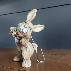 Könnte beinhalten: Eine beige Kaninchenfigur, die auf den Hinterbeinen steht und einen grün-rot gestreiften Schal trägt. Das Kaninchen hält eine kleine braune Geige und einen Bogen, wobei ein dünner silberner Draht von der Seite des Instruments zum Sockel führt.
