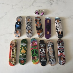 Puede incluir: Una colección de once monopatines para dedos con varios diseños gráficos, incluyendo una calavera, un símbolo de paz, una corona y patrones abstractos, junto con dos llaveros de metal plateado con pequeñas anillas de goma moradas.