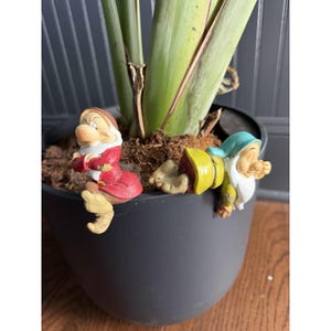 以下が含まれることがあります： ダークグレーの植木鉢の縁に置かれた2つの小さな装飾用フィギュア。1つは赤いコートと茶色の帽子をかぶり、不機嫌そうな表情をしています。もう1つは黄色いシャツと青緑色の帽子をかぶり、横たわって眠そうな表情をしています。