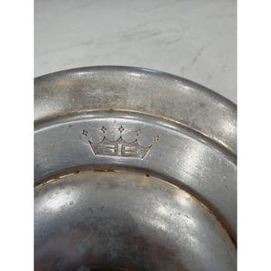 Puede incluir: Un plato de color plateado con un diseño decorativo de corona y diamantes. El plato tiene una forma ligeramente cóncava y una superficie lisa y reflectante. El diseño de la corona está grabado en el metal.