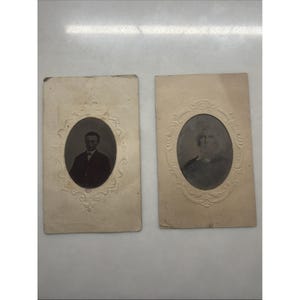 May include: Two rectangular off-white paper frames with embossed oval borders, each containing a black and white portrait photograph. The left frame features a person in a dark suit, while the right frame displays a person with light hair.
