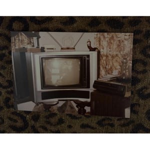 May include: A vintage photograph of a television set with a white cabinet and a dark screen. The TV sits on a table with other items, including a radio antenna, a decorative object, and a cassette player. The background features patterned curtains.