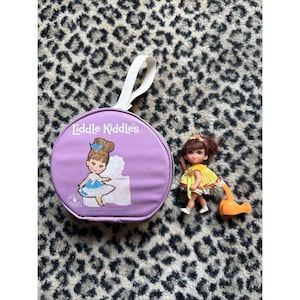 Könnte beinhalten: Ein Vintage-Liddle-Kiddles-Puppen-Set. Es enthält eine runde, lila Tasche mit weißem Griff, eine Puppe in einem gelben Kleid und ein orangefarbenes Zubehör. Auf der Tasche steht "Liddle Kiddles" und eine Puppenzeichnung.
