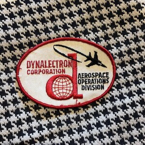 May include: An oval patch with the text "Dynalectron Corporation Aerospace Operations Division" in red and black. The patch features a stylized globe and a jet plane. The background is a black and white houndstooth pattern.