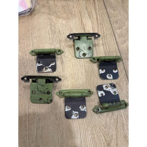 May include: Five metal cabinet hinges with a distressed finish, featuring a sage green top section and a black bottom section. Each hinge has a cylindrical barrel and mounting holes, showing visible paint wear and chipped surfaces on the metal.