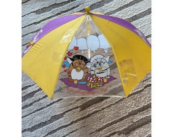 Puede incluir: Un paraguas vintage con una capota amarilla y morada. El paraguas presenta un panel transparente con una ilustración de dibujos animados de dos personajes Furby haciendo un picnic. El texto "Kah-may-may-u-nye" está impreso en el paraguas.