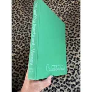 Puede incluir: Un libro de tapa dura verde brillante titulado "You and Your Apartment - Varney". El libro tiene texto blanco en el lomo y una firma en la portada. También es visible el nombre de la editorial, Bobbs-Merrill.