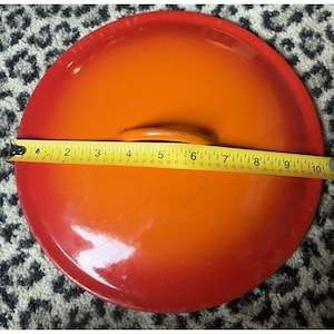 May include: A round, enameled cast iron pot lid with a red to orange gradient. The lid is approximately 10 inches in diameter. A yellow measuring tape is laid across the lid, showing the diameter measurement.