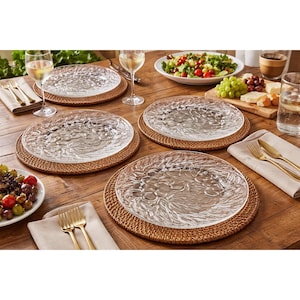May include: A table setting with clear glass plates featuring a grape design, placed on woven brown placemats. Gold-colored forks and knives are next to beige napkins. Wine glasses and food items complete the dining arrangement.