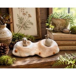 May include: A light-colored, textured ceramic sculpture featuring two small, sculpted animal figures nestled in rounded indentations. The piece has a smooth, matte finish with subtle brown accents and small, raised circular details along the base.