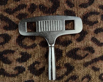 Llave de cuerda para reloj antigua, grande, de tono plateado, con orificio cuadrado.