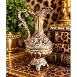 May include: A gold-toned decorative pitcher with a distressed finish, featuring an ornate handle, a fluted neck, and a bulbous body adorned with embossed floral motifs. The vessel stands on a pedestal base with three small feet and a scalloped rim.
