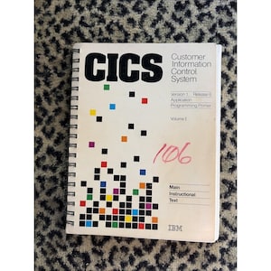 Puede incluir: Un libro encuadernado en espiral titulado "CICS Customer Information Control System" con el texto "Version 1 Release 6 Application Programming Primer Volume I." La portada presenta un diseño pixelado colorido y el número "106" escrito en rojo.